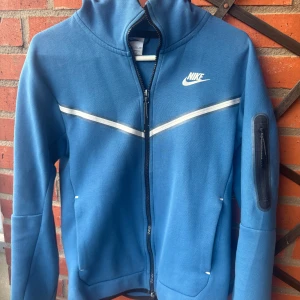 Blå Nike tech fleece - Säljer en blå hoodie från Nike med dragkedja och vit logga på bröstet. Tröjan har en hög krage och en praktisk ficka med dragkedja på ärmen. Perfekt för en sportig look!