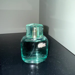Grön parfymflaska från Lacoste - Säljer en stilren grön parfymflaska från Lacoste. Flaskan har en elegant och kompakt design med det ikoniska krokodilmärket på framsidan. Perfekt för den som vill ha en fräsch och sportig doft.