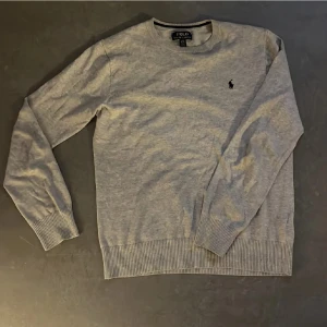 Grå tröja från Polo Ralph Lauren - Säljer en stilren grå tröja från Polo Ralph Lauren med det klassiska broderade logotypen på bröstet. Tröjan har långa ärmar och ribbstickade muddar vid ärmslut och nederkant. Perfekt för en avslappnad och elegant look.storlek XL i barn storlek som motsvarar xs/s