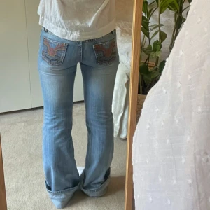 Ljusblå bootcut jeans med unika fickor - Snygga ljusblå bootcut jeans med unika broderade detaljer på bakfickorna. Jeansen har en låg midja och en klassisk femficksdesign.  Jeansen har ingen stretch och är ca 8 dm dör långa på mig som är 160 (dock snyggt att dom är långa). Skriv om du har frågor!💕