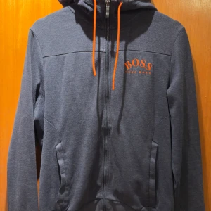 Mörkblå hoodie ”Saggy” från Hugo Boss (S) - Snygg mörkblå hoodie från Hugo Boss med orange detaljer. Tröjan har en dragkedja framtill och justerbar huva med snören. Perfekt för en avslappnad stil. 