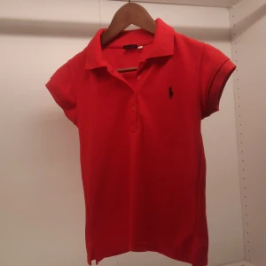 Röd barn pikétröja från Polo Ralph Lauren - Säljer en klassisk röd pikétröja från Polo Ralph Lauren med korta ärmar och den ikoniska loggan broderad på bröstet. Perfekt för en stilren och avslappnad look. Tröjan har en krage och knappar framtill.