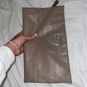 Beige kuvertväska med krokodilmönster - Snygg beige kuvertväska med krokodilmönster och ett stilrent G-emblem. Väskan har en praktisk dragkedja och ett handtag på baksidan för enkel bärning. Insidan är fodrad med ett mönster av svarta G-bokstäver på vit bakgrund.
