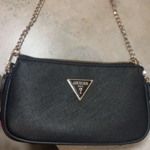 Svart axelväska från Guess - Snygg svart axelväska från Guess med en kedjerem och dragkedja. Väskan har en stilren design med det ikoniska Guess-logotypen i metall på framsidan. Perfekt för att ge din outfit en elegant touch.