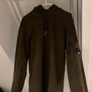 Olivgrön hoodie från C.P. Company - Säljer en snygg olivgrön hoodie från C.P. Company. Den har en cool detalj med en lins på ärmen och en justerbar huva. Perfekt för en avslappnad stil.