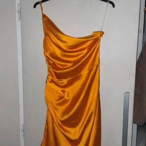 Orange satin klänning - Snygg orange klänning i satin med en axelbandslös design. Klänningen har en draperad stil som ger en elegant look. Perfekt för en kväll ute eller en speciell tillställning.