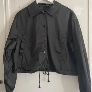 Svart jacka från H&M - Snygg svart jacka från H&M med knappar och dragsko i nederkant. Perfekt för en stilren look. Jackan har långa ärmar och en klassisk krage.