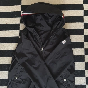 Svart vindjacka från Moncler - Snygg svart vindjacka från Moncler med huva och dragkedja. Jackan har Moncler-logga på bröstet och en stilren design med knappar på fickorna. Perfekt för blåsiga dagar.