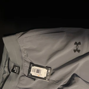 Grå träningsbyxor från Under Armour - Snygga grå träningsbyxor från Under Armour med elastisk midja och logotyp på benet. Perfekta för träning med en bekväm passform och stilren design. Byxorna har en rak stil och är tillverkade i ett syntetmaterial som andas.