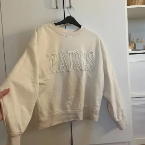 Beige sweatshirt från H&M med text - Säljer en beige sweatshirt från H&M med texten 'PARIS' i stora bokstäver på framsidan. Tröjan har en avslappnad passform och långa ärmar, perfekt för en casual look.