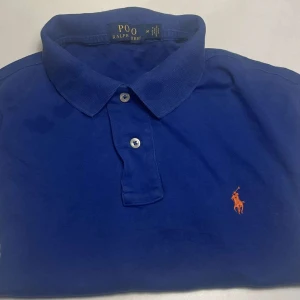 Blå pikétröja från Ralph Lauren - Säljer en klassisk blå pikétröja från Ralph Lauren med den ikoniska loggan broderad i orange på bröstet. Tröjan har en krage och knappar framtill, perfekt för en stilren look. Passar bra till både jeans och chinos. 