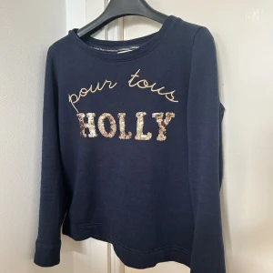 Mörkblå tröja från Holly & Whyte - Säljer en mörkblå tröja från Holly & Whyte med texten 'pour tous HOLLY' i glittriga bokstäver på framsidan. Tröjan har långa ärmar och en rund halsringning. Perfekt för en avslappnad stil.