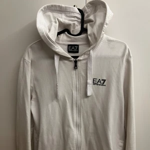 Vit hoodie ea7 - Säljer en stilren vit hoodie från Emporio Armani med dragkedja och huva. Den har en diskret logga på bröstet och snören i huvan. Perfekt för en avslappnad look.