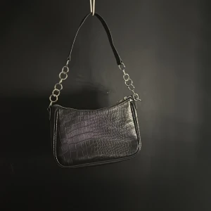 Svart axelväska med krokodilpräglat mönster - Snygg svart axelväska med krokodilpräglat mönster och stiliga metallringar på axelremmen. Väskan har en dragkedja för säker förvaring och en elegant look som passar perfekt för olika tillfällen.