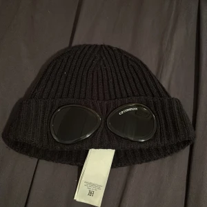 C.P. Company mössa - Snygg svart stickad mössa från C.P. Company med unika glasögondetaljer framtill. Perfekt för att ge din outfit en cool touch. Mössan är ribbad och har en klassisk passform. 8/10 skick väldigt bra.väldigt mörk blå näst intill svart. Kvitto finns om man vill se de men skriv inte om ni är halv säkra på ett köp. Skam bud svaras ej. Hör av dig vid intresse.