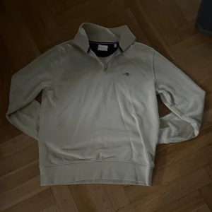 Beige tröja från GANT - Säljer en  beige half zip tröja från GANT med dragkedja vid halsen och broderad logotyp på bröstet. Tröjan har långa ärmar och är tillverkad i ett mjukt material.