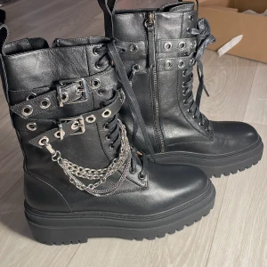 Svarta boots med kedjedetaljer från Zara - Snygga svarta boots från Zara i skinn med coola kedjedetaljer och snörning. De har en robust sula och dragkedja på sidan för enkel på- och avtagning. Perfekta för en edgy look!