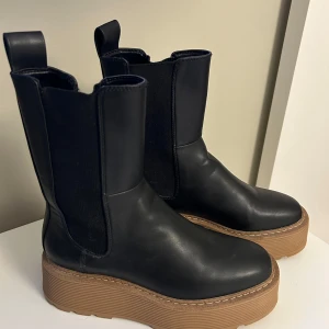 Svarta platåstövletter - Säljer ett par snygga svarta platåstövletter i skinn. De har en rejäl brun sula som ger extra höjd och stil. Perfekta för höst och vinter med sin robusta design. Skorna är i nyskick och redo att användas för att lyfta din outfit!