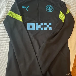 Svart långärmad tröja från Puma - Säljer en svart långärmad tröja från Puma med coola detaljer i blått och gult. Tröjan har en dragkedja framtill och är perfekt för träning eller vardagsbruk. Den är i bra skick och har en snygg passform. Perfekt för dig som gillar sportig stil! Manchester city 