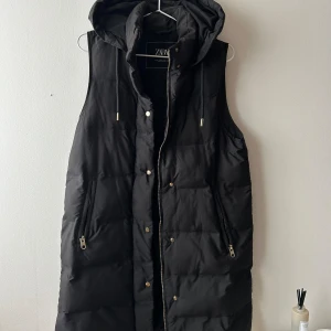 Zara väst - Lång svart väst från Zara med gulddetaljer. Luva, dragkedja, gulddetaljer, svart, väst, bra skick. Storlek S. Köpt för 699kr förra året. Använd Max 4 ggr.