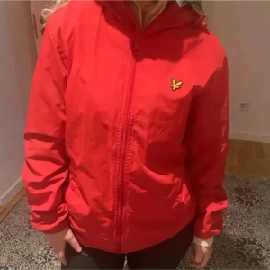 Röd vindjacka från Lyle & Scott - Säljer en snygg röd vindjacka från Lyle & Scott. Jackan har en dragkedja framtill och en huva för extra skydd mot väder och vind. Perfekt för vår och höst!