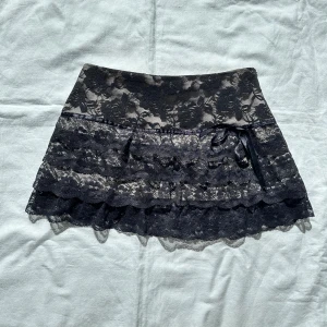 Svart spetskjol med volanger - Säljer en superfin svart kortkjol i spets med volanger. Kjolen har ett snyggt blommigt mönster och en söt rosett på sidan. Perfekt för fest eller en utekväll!Vid köp av flera varor får du rabatt 💛