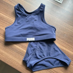 Mörkblå bikini från Puma - Säljer en snygg och sportig mörkblå bikini från Puma. Toppen har en hög halsringning och breda axelband för extra stöd, perfekt för både bad och träning. Bikinitrosorna har en klassisk passform. Materialet är mjukt och stretchigt för en bekväm passform. Puma-loggan syns diskret på både topp och trosor. Tyvärr hann jag klippa bort lappen med storleken innan min dotter provade den, den passar 9-10 åring. 