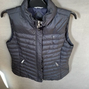 Svart dunväst från GANT - Säljer en snygg och praktisk svart dunväst från GANT. Västen har en quiltad design med dragkedja och knappar framtill. Den är perfekt för höst och vinter och har två praktiska fickor med dragkedjor. Passar både till vardags och lite mer uppklädda tillfällen.