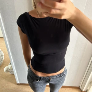 Svart croppad topp med öppen rygg - Säljer en snygg svart croppad topp med en cool öppen ryggdesign. Toppen är ärmlös och har en tight passform, perfekt för fest eller en utekväll. Den är gjord i ett mjukt och stretchigt material som sitter skönt på kroppen.