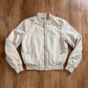 Beige bomberjacka från stradivarius - Säljer en snygg beige bomberjacka i storlek M. Jackan är tillverkad i 100% polyester och har en stilren design med dragkedja framtill. Den har långa ärmar och ribbade muddar vid ärmslut och nederkant. Perfekt för vår och höst!