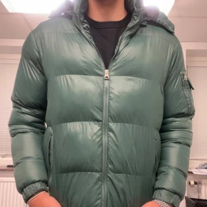 Grön pufferjacka från Brave Soul - Säljer en grön pufferjacka från Brave Soul i storlek XL. Jackan har en dragkedja framtill och är långärmad med en luva. Den har också praktiska fickor med dragkedjor för säker förvaring. Passar perfekt för vintern!