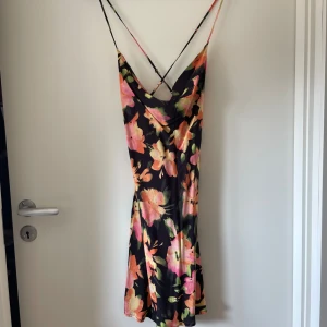 Blommig sommarklänning med öppen rygg - Säljer en superfin blommig klänning perfekt för sommaren! Den har en öppen rygg med tunna axelband och ett färgglatt blommönster i rosa, orange och grönt på en svart bakgrund. Klänningen är i ett lätt och luftigt material, perfekt för varma dagar.