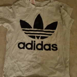 Vit t-shirt från Adidas - Säljer en vit t-shirt från Adidas med det klassiska svarta logotyptrycket på framsidan. Perfekt för en sportig look eller avslappnad stil.