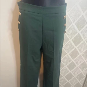Gröna byxor med guldknappar L storlek  - Snygga gröna byxor med hög midja och dekorativa guldknappar på sidorna. Byxorna har en rak passform och är perfekta för en stilren look.