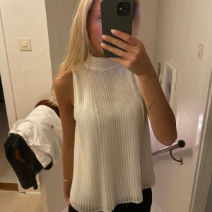 Vit ärmlös topp - Säljer en stilren vit ärmlös topp med hög hals och plisserad design. Perfekt för en elegant och luftig look.