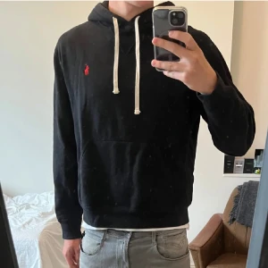 Svart hoodie från Ralph Lauren - Säljer en snygg svart hoodie från Ralph Lauren med klassisk röd logga på bröstet. Den har en justerbar huva med vita snören och en praktisk magficka. Perfekt för en avslappnad stil eller kyliga dagar. Passar till det mesta och är superbekväm!