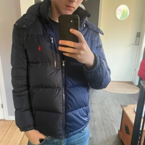 Mörkblå dunjacka från Ralph Lauren - Säljer en snygg och varm mörkblå dunjacka från Ralph Lauren. Jackan har en huva och dragkedja framtill, perfekt för kalla vinterdagar. Den är i väldigt br skick. Passar perfekt för både vardag och lite finare tillfällen.  Storleken är Xl ungdom passar xs-S