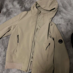 Beige jacka från C.P. Company - Säljer en snygg beige jacka från C.P. Company i storlek M. Jackan är tillverkad i C.P. Shell-R material, vilket är ett mjukt och stretchigt softshell-tyg av återvunnet garn. Den har en dragkedja framtill och praktiska fickor med dragkedjor. Perfekt för höst och vår! Nypris ligger den på 5576kr