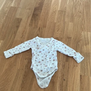 Vit blommig body från Newbie - Söt vit body från Newbie med blommigt mönster i rosa och grönt. Bodyn har långa ärmar och praktiska tryckknappar i grenen för enkel på- och avklädning. Perfekt för de allra minsta!