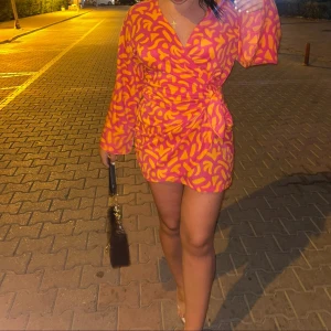 Färgglad omlottklänning - Säljer en färgglad omlottklänning med ett livligt mönster i orange och rosa. Klänningen har långa ärmar och en knytning i midjan för en smickrande passform. Perfekt för att lysa upp garderoben! 