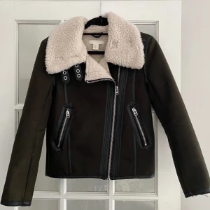 Mörk grön  jacka med fuskpäls - Säljer en snygg mörk grön jacka med fuskpäls på kragen. Den har dragkedja och coola detaljer med knappar. Perfekt för höst och vinter, håller dig varm och stilren.
