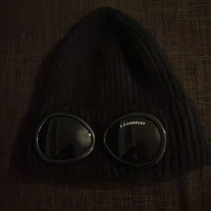  mössa C.P. Company - Säljer en cool svart ribbad mössa från C.P. Company med inbyggda glasögon. Perfekt för att sticka ut i mängden och hålla sig varm samtidigt. Passar både till vardags och speciella tillfällen. Ett måste för den stilmedvetna!