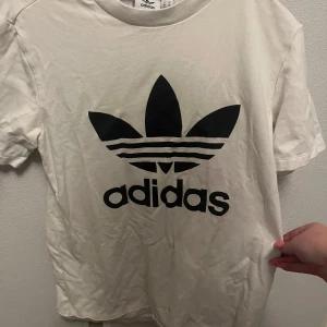 Vit t-shirt från Adidas - Säljer en klassisk vit t-shirt från Adidas med deras ikoniska logga i svart på framsidan. T-shirten är i bomull och har en normal passform med korta ärmar. 