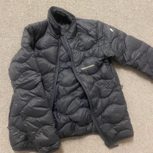 Marinblå dunjacka från Peak Performance  - Säljer en mörkblå dunjacka från Peak Performance med quiltad design och dragkedja. Perfekt för kyliga dagar med sin varma och lätta konstruktion. Jackan har en hög krage och långa ärmar för extra skydd mot kylan. Storlek 150 barn men passar xxs/xs, finns en skada kolla de två sista bilderna