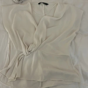 Vit omlottblus från Zara - Säljer en elegant vit omlottblus från Zara. Blusen har en knytning i midjan och är ärmlös, vilket ger en stilren look. Perfekt för både vardag och festliga tillfällen.