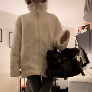 Vit teddyjacka med dragkedja - Mysig vit teddyjacka med dragkedja och hög krage. Perfekt för kyliga dagar med sin fluffiga och varma design. Jackan har långa ärmar och två praktiska fickor framtill.