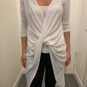 Lång topp  från Flam Mode Ny - Säljer en elegant vit lång topp från Flam Mode. Den har en omlott-design med draperade detaljer framtill och trekvartsärmar. Perfekt för sommarens fester eller en stilren vardagslook. Materialet är lätt och luftigt, vilket gör den bekväm att bära. Storlek M-L