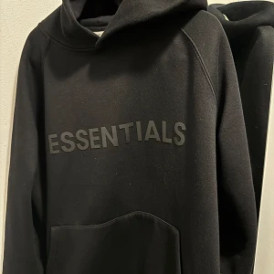 Svart hoodie från Essentials - Säljer en stilren svart hoodie från Essentials med logga på bröstet. Perfekt för en avslappnad look. Den har en klassisk huva och en stor ficka framtill.
