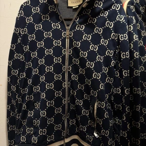 Mönstrad tröja från Gucci - Säljer en snygg blå tröjan från Gucci med det ikoniska GG-mönstret. Tröjan har dragkedja och långa ärmar, samt en randig detalj i rött och vitt längs ärmarna. Perfekt för dig som vill ha en stilren och lyxig look. ”Storlek L passar bra som M också”