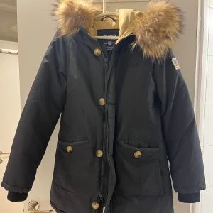 Svart parkajacka från Svea - Säljer en snygg svart jacka från Svea i storlek XS. Jackan har en mysig pälskantad huva och stängs med både dragkedja och knappar. Perfekt för kalla vinterdagar!  